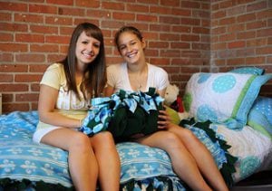 Two college roommates meeting for the first time and sitting in a bed inside a dorm room.